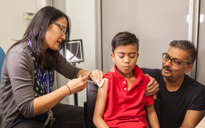 BCCH patient, Hans takes a breath as a clinician gives him an immunization shot with his dad kneeling beside him